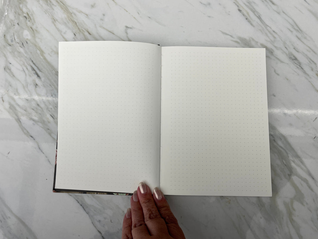 JL-0086E - Skulls &amp; Flowers Pattern Dot Grid Journal - Open dot grid journal with cream pages and dark patterned spine, held by hand against marble surface