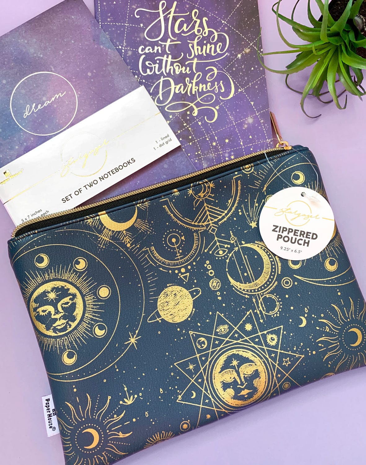 Navy pencil pouch with gold zipper featuring gold celestial illustrations shown on a purple background with a set of two purple notebooks.