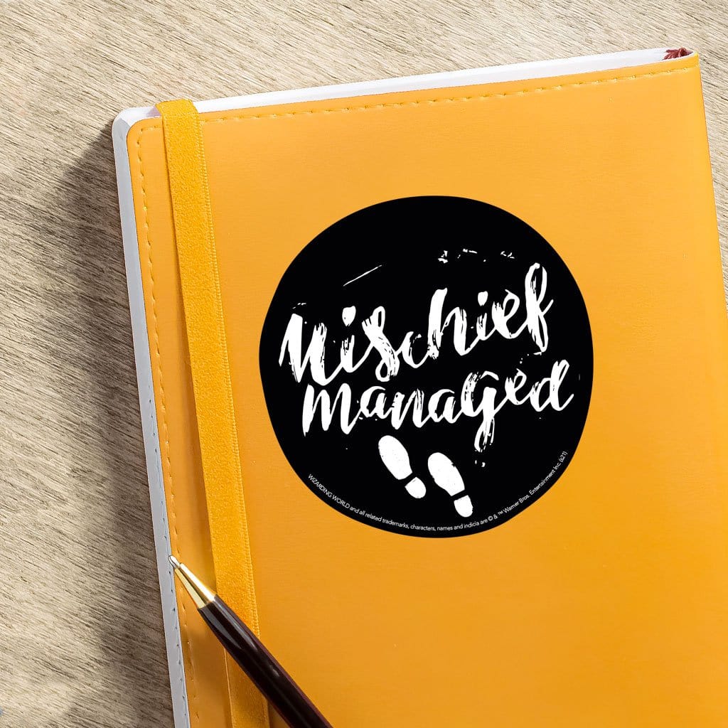 vinyl laptop sticker featuring Harry Potter Mischief Managed on black, circle background shown adhered to orange notebook with pen.